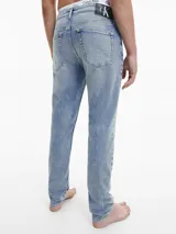 Jeans de mezclilla azul claro, tiro medio, con corte slim que se estrecha hacia el tobillo. Presentan un diseño de cinco bolsillos, bragueta con cremallera y herrajes de Calvin Klein, incluyendo una insignia de cuero en la parte trasera.