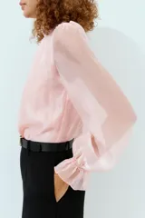 Mono largo con parte superior de blusa rosa de manga larga, cuello alto fruncido y puños con volantes. La parte inferior es un pantalón negro de pernera ancha con bolsillos delanteros y un cinturón negro con hebilla dorada.