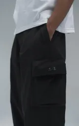 Pantalón cargo de estilo técnico en color negro, confeccionado en textil memory con acabado sutilmente brillante. Presenta una silueta oversize, cinturón regulable integrado con hebilla de encastre, dos bolsillos laterales, dos bolsillos cargo de gran tamaño y un bolsillo trasero.
