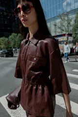 Chaqueta efecto piel color marrón, con cuello solapa, mangas por debajo del codo, cintura elástica, falsos bolsillos delanteros y cierre frontal con cremallera.