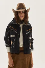 Campera de lana merino hervida en color crudo, con diseño de cuello camisero, cierre frontal de botones y bolsillos frontales. Presenta bordados artesanales con motivos geométricos en tonos marrón y azul en el frente, espalda y mangas.