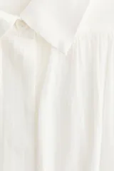 Blusa oversize blanca de algodón y viscosa, con cuello camisero, tapeta de botones ocultos y detalles fruncidos en el canesú delantero y trasero. Presenta mangas globo largas con puños abotonados.