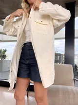 Camisa color crema de corte amplio, con cuello clásico, cierre frontal con botones a presión, mangas largas con puños abotonados y bolsillos de parche en el pecho.