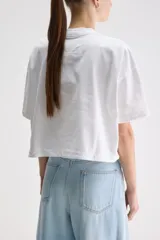 Remera cropped blanca de calce relajado, con cuello redondo, hombros caídos y mangas cortas. Presenta un pequeño detalle de etiqueta bordada en el lateral inferior.
