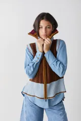 Chaleco de lana con cierre frontal completo y cuello alto. Presenta un diseño de bloques de color en tonos marrón chocolate y celeste, combinando secciones de tejido trenzado con secciones de tejido acanalado. Posee un corte asimétrico, siendo más largo en la parte posterior, y aberturas laterales en el ruedo.
