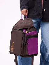 Mochila Rusty de lona color marrón con detalles en violeta. Presenta un compartimento principal con solapa, bolsillo frontal con cierre y panel de malla violeta, y logo estampado en el lateral.