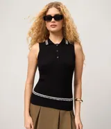 Blusa sin mangas de punto color negro, con cuello polo y cierre frontal de tres botones. Presenta detalles de líneas blancas contrastantes en el cuello y en el ruedo.