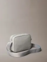 Cartera tipo cámara gris claro de poliuretano texturizado, con cierre superior de cremallera, correa de hombro ajustable y monograma CK grabado en la esquina delantera.