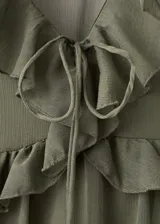 Vestido midi color verde militar, confeccionado en tejido fluido de poliéster. Presenta un diseño evasé, escote de pico con volantes, y mangas largas con puños elásticos. Incluye detalles de volantes en el cuello y forro interior.