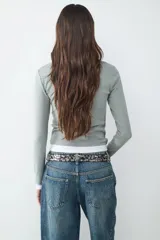 Camiseta de manga larga con diseño de doble capa, confeccionada en jersey suave y elástico. Presenta un calce ajustado, cuello redondo y un estilo minimalista que combina una capa superior en color verde grisáceo con una capa inferior en color blanco.