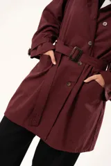 Trench de corte holgado color bordó, con cuello camisero, cierre frontal con botones y cinturón ajustable con hebilla. Presenta mangas largas con detalles de correas en los puños y bolsillos laterales.
