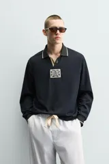 Polo de manga larga en tejido técnico acanalado color negro, con cuello solapa y abertura frontal. Presenta puños con ribete a contraste y una etiqueta aplicada en el pecho con el diseño conmemorativo de la Copa Mundial de la FIFA 1966.