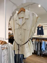 Camisa de lino color beige, de silueta relajada, estilo utility, con cuello camisero, mangas cortas caídas, bolsillos frontales con solapa, cierre de botones y cinturón delgado trenzado marrón.