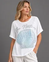 Remera blanca de corte holgado con mangas cortas con dobladillo. Presenta un estampado frontal con la frase 'The Nature' en letras plateadas brillantes y un aplique central de una caracola en tonos tornasol decorada con pequeñas perlas.