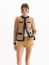 Chaqueta entallada de twill color marrón con detalles en negro que ribetean el cuello redondo, la línea de botones, el ruedo, los puños y los bolsillos. Presenta broches dorados en el frente y las mangas.