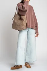 Cartera tipo shopper de gamuza sintética color marrón, con diseño estructurado, asa de hombro ajustable y detalle de placa metálica dorada con logo en la parte frontal.