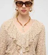 Blusa tipo bata de encaje texturizado color beige, con escote en V, mangas largas y volados aplicados en el escote. Presenta cierre frontal con botones y lazo de ajuste en la cintura.