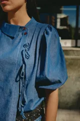 Blusa de denim liviano de corte asimétrico en tono azul, con mangas abullonadas, cuello redondo y cierre lateral con lazos para atar y botones.