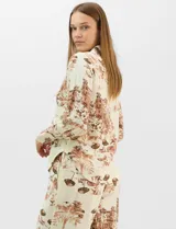 Conjunto de dos piezas compuesto por camisa de manga larga con cuello solapa y botones frontales, y pantalón de tiro alto con pierna recta y bolsillos laterales. Ambas prendas presentan un estampado paisajístico en tonos marrones sobre fondo crema.