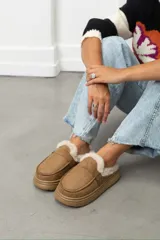 Zueco de gamuza color beige con interior forrado en piel sintética, suela de plataforma plana y detalle de hebilla lateral.