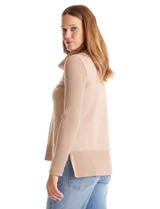 Buzo de punto color beige con cuello volcado amplio, mangas largas acanaladas y torso liso. Presenta una costura central vertical, ruedo con tajos laterales y largo asimétrico, siendo más largo en la parte posterior.
