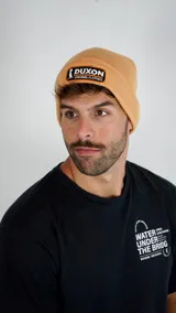Gorro tipo beanie color tostado, con parche rectangular frontal con el logo de la marca Duxon bordado en blanco y naranja.