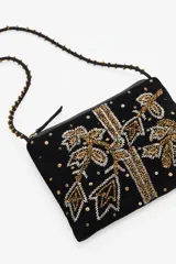Cartera tipo bandolera de terciopelo negro con bordados artesanales en pedrería dorada y plateada. Cuenta con una correa larga decorada con cuentas y borlas en los extremos, cierre superior y forro interior de algodón.