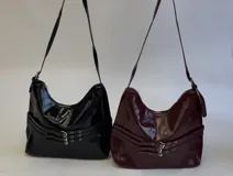 Cartera bandolera negra de efecto charol, con diseño hobo y tres tiras decorativas horizontales con hebillas plateadas en la parte inferior. Cuenta con correa larga regulable y cierre superior.