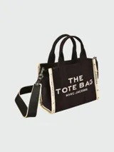 Cartera tipo tote bag de lona negra con letras blancas estampadas en el frente. Cuenta con doble asa superior, correa ajustable y desmontable, y detalles laterales en contraste.