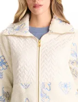 Chaqueta de corte relajado confeccionada en tejido tipo corderito color crema, combinada con paneles de matelassé en el frente y bolsillos. Presenta un delicado bordado floral en tonos azules sobre el corderito, cierre frontal metálico dorado y bolsillos de parche delanteros.