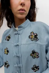Chaqueta liviana de jean celeste con cuello mao, cierre frontal con alamares y detalle de bordados florales.