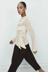 Jersey de punto color crudo con cuello redondo y manga larga. Presenta un diseño calado con detalles de encaje en el frente y un bajo asimétrico.