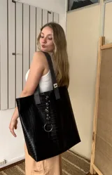 Tote bag de gran tamaño confeccionada en ecocuero negro con textura de reptil. Presenta una tira central decorativa con ojales metálicos y una argolla grande en la parte inferior, además de manijas en color gris.