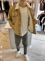 Chaqueta de pana color marrón tostado, con forro interno y cuello de corderito sintético blanco. Presenta cierre frontal de botones metálicos, bolsillos frontales con solapa en el pecho y bolsillos laterales.