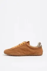 Zapatilla deportiva de estilo casual confeccionada en serraje vacuno color marrón. Presenta un diseño con empeine festoneado, cordones en tono contrastante, detalle de piel en el talón y suela de caucho con relieve. Incluye una cuña interior para una ligera elevación.