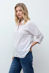 Camisa de corte holgado con diseño de rayas verticales en tonos beige y blanco, con sutiles destellos brillantes. Presenta cuello clásico, cierre frontal con botones y mangas largas con puños ajustables.