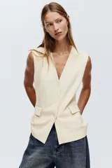 Chaleco largo color beige, con escote en V, cierre de botones a presión ocultos y bolsillos decorativos frontales. Presenta cintura entallada con pinzas y trabilla con botón en la espalda.