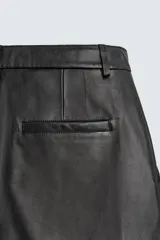 Bermuda de corte relajado confeccionada en piel negra, con pliegues frontales, bolsillos laterales y cierre frontal con cremallera y botón.