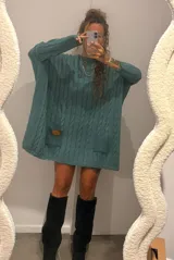 Sweater de punto color camel con diseño de trenzas verticales y acabado brillante. Presenta una silueta holgada tipo poncho, cuello redondo, mangas largas con puños ajustados y bolsillos frontales con detalle de etiqueta de cuero.