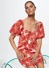 Vestido corto de algodón con estampado de margaritas rojas sobre fondo blanco. Presenta un diseño evasé, escote cuadrado con cierre de lazo ajustable, mangas cortas abullonadas y forro interior.