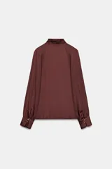 Blusa de tejido satinado color bordó, con cuello subido drapeado y manga larga con puños. Presenta detalle de pliegues frontales y cierre con botones laterales en el cuello.