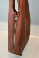 Bolso tipo tote de gran capacidad, confeccionado artesanalmente en cuero vacuno color marrón. Presenta un diseño minimalista con costura central vertical, fuelle lateral ajustable con cierre y placa metálica con logo en la parte inferior.