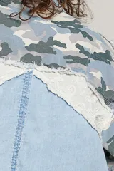 Chaqueta de denim de corte holgado y manga larga, con diseño en técnica de patchwork que combina sectores de mezclilla clásica, estampa de camuflaje, parches florales y detalles de encaje blanco. Cuenta con cierre frontal de cremallera metálica y cuello camisero.