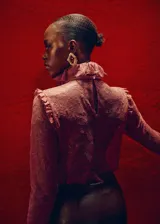 Blusa de encaje color bordó con cuello gorguera, lazo frontal y mangas largas con volantes en los puños. Presenta un diseño recto con cierre de cremallera en la parte posterior e incluye forro interior.