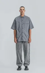 Camisa de manga corta de poplin, color gris, con silueta oversize. Presenta dos bolsillos delanteros, uno con solapa y otro con ojal y botón.