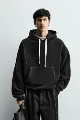 Sudadera tipo hoodie de corte relajado, color negro, con interior perchado. Presenta capucha ajustable con cordones en color crudo, bolsillo frontal tipo canguro y costuras a contraste en color blanco. Posee puños y ruedo en rib.