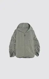 Campera rompevientos de diseño oversize, color verde, confeccionada en microfibra técnica ligera. Presenta capucha integrada, cierre frontal técnico y un sistema de frunces elásticos regulables a lo largo de las mangas que permiten ajustar el volumen de la prenda.