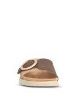 Sandalias tipo slide con tira ancha en tejido texturizado color beige y detalle de hebilla circular decorativa en tono marrón. Presentan una base ergonómica con suela de poliuretano ligera y flexible.