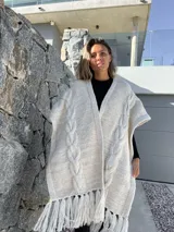 Ruana tejida en lana merino, color beige y crudo, con diseño de trenzas verticales en el frente y terminación con flecos en el ruedo.