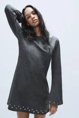Vestido corto de punto color gris oscuro, con cuello barco y mangas largas acampanadas. Presenta detalles de tachas metálicas aplicadas en el ruedo y en los puños, con un diseño de corte evasé.